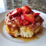 A single, gooey Strawberry Shortcake Cinnamon Roll topped with sliced strawberries and glaze, with a bite taken out.