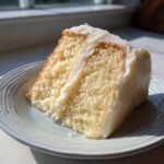 A close-up of a moist slice of Vanilla Birthday Cake with thick white buttercream frosting on a light blue plate.