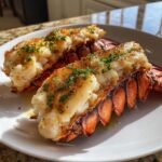 Two succulent Baked Lobster Tails, split open and topped with butter and fresh parsley, served on a white plate.