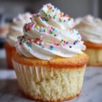 A close-up of one perfect vanilla cupcake with white frosting and colorful sprinkles, part of a batch of The Best Vanilla Cupcakes With Sprinkles.