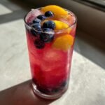 Close-up of vibrant red Blueberry Peach Lemonade garnished with fresh blueberries and peach slices.