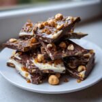 A stack of broken pieces of Cashew And Hazelnut Toffee Chocolate Bark topped with whole hazelnuts and amber toffee bits.