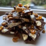 Pieces of Cashew And Hazelnut Toffee Chocolate Bark stacked on a white plate, featuring dark chocolate, nuts, and amber toffee shards.