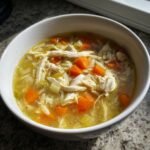 Close-up of a white bowl filled with steaming Chicken Orzo Soup, featuring shredded chicken, bright orange carrots, and orzo pasta.