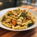 A close-up of a serving of Chicken Pasta Primavera featuring penne pasta, seasoned chicken chunks, zucchini, broccoli, and carrots.