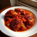 A white plate filled with rich, rustic Chicken Cacciatore featuring chicken pieces simmered in a thick tomato sauce with carrots and celery.