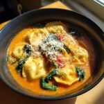 A close-up of a dark bowl filled with Cozy Tuscan Ravioli Soup, featuring cheese ravioli, spinach, and grated Parmesan.