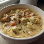 A close-up of a white bowl filled with Creamy Blackened Chicken Tortellini Soup, showing pasta, chicken chunks, and carrots.