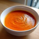 A close-up of a white bowl filled with vibrant orange Creamy Roasted Red Pepper Tomato Soup, garnished with pepper.