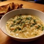 A close-up of a white bowl filled with Creamy White Bean Lemon Orzo Soup, garnished with fresh parsley.
