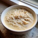 A close-up of a white bowl filled with thick, creamy white chicken rice soup, featuring shredded chicken and white rice.