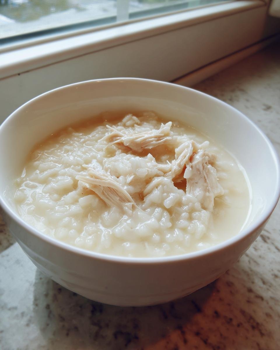 Close-up of a white bowl filled with creamy white chicken rice soup, topped with shredded chicken.