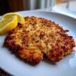 Close-up of a perfectly golden and crispy Chicken Milanese served on a white plate with two lemon wedges.