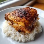 A close-up of a crispy, seasoned chicken thigh resting on a bed of fluffy white rice, showcasing our Chicken Thighs With Rice recipe.