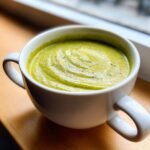 A close-up of a white mug filled with thick, green Delicious Cream Of Broccoli Soup, topped with cracked pepper.
