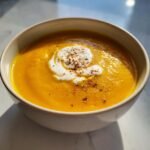 A close-up of a bowl of vibrant orange Easy Roasted Butternut Squash Soup topped with a swirl of cream and cracked pepper.