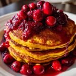 A tall stack of fluffy Pumpkin Pancakes With Cranberry Maple Syrup, topped with whole cranberries.