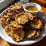 A plate piled high with golden brown Fried Zucchini Chips served with a small ramekin of garlic vinaigrette.