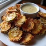 A plate piled high with golden brown Fried Zucchini Chips served with a small cup of creamy garlic vinaigrette.