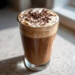 A tall, frosty glass filled with a Frozen Chocolate Brandy Alexander, topped with whipped cream and chocolate shavings.