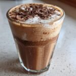 A tall glass filled with a creamy, frothy Frozen Chocolate Brandy Alexander, topped with chocolate shavings.