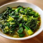 A close-up of a white bowl filled with vibrant green vegetables, including broccoli florets and leafy greens, submerged in a savory broth of the Immune Boosting Green Veggie Swamp Soup.