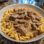 A close-up of a white bowl filled with Irresistible Beef Stroganoff served over wide egg noodles, featuring chunks of beef and sliced mushrooms in a creamy sauce.