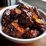 A close-up of glossy, dark pieces of Irresistible Black Pepper Chicken coated in a rich sauce with visible black pepper flakes and onion slices.