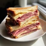 Two halves of an Irresistible Roast Beef And Cheese Sandwiches stacked on a white plate, showing layers of melted cheese and thinly sliced roast beef.