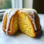 A slice cut from a small Lemon Ginger Bundt Cake showing the moist yellow crumb, topped with white glaze and lemon zest.