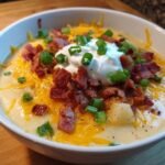 A close-up of a bowl of creamy Loaded Baked Potato Soup topped generously with shredded cheddar, bacon bits, sour cream, and green onions.