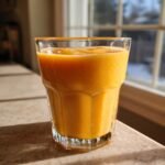 A vibrant, thick Mango Banana Smoothie served in a clear, textured glass, backlit by sunlight near a window.