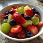A close-up of a vibrant Moscato Fruit Salad featuring strawberries, blueberries, green grapes, and pineapple chunks in a white bowl.