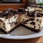 A rich slice of No Bake Cookies And Cream Cheesecake with a dark chocolate crust and creamy filling studded with cookie pieces.