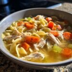 Close-up of a bowl of Nourishing Chicken Noodle Soup featuring shredded chicken, egg noodles, and bright orange carrots.