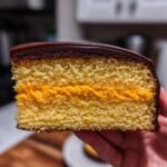 Close-up of a slice showing the layers of Orange Buttercream Filled Jaffa Cakes: sponge, orange filling, and chocolate topping.