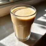 A tall glass filled with a creamy, light brown Peanut Butter Banana Smoothie, sitting on a counter in bright sunlight.