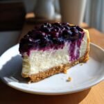 A rich slice of Perfect Lemon Blueberry Cheesecake with a thick blueberry topping on a white plate.