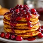 A tall stack of fluffy Pumpkin Pancakes With Cranberry Maple Syrup, topped with rich cranberry compote and fresh berries.