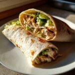 Two halves of a freshly made Chicken Caesar Wrap filled with shredded chicken, lettuce, and creamy dressing, resting on a white plate.