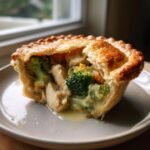A single serving of Roasted Broccoli Cheddar Chicken Pot Pie with a flaky golden crust broken open to show creamy filling, chicken, and bright green broccoli.