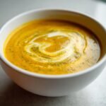 Close-up of rich, orange Roasted Broccoli Cheddar Soup swirled with cream and spices in a white bowl.