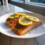 Two fillets of bright orange Slow Cooker Salmon garnished with fresh dill and thin lemon slices on a white plate.