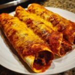 Three rolled Spicy Black Bean Enchiladas covered in rich red sauce and melted cheddar cheese on a white plate.