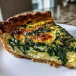 A close-up of a thick slice of Spinach Quiche showing the rich, dark green filling and golden-brown crust on a white plate.