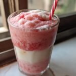 Close-up of a layered Strawberry Cheesecake Protein Shake with pink and white sections and a red and white striped straw.