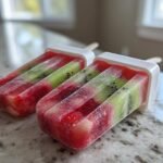 Two homemade Strawberry Kiwi Fruit Popsicles, layered with red strawberry pieces and green kiwi slices, resting on a light countertop.