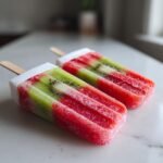 Two layered Strawberry Kiwi Fruit Popsicles, red and green, resting on a white marble surface.
