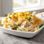 A close-up of a white bowl filled with Turkey Legs Rice Casserole, topped with melted cheddar and green peas.