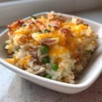 A close-up of a single serving of Turkey Legs Rice Casserole topped with melted, browned cheddar cheese and peas.